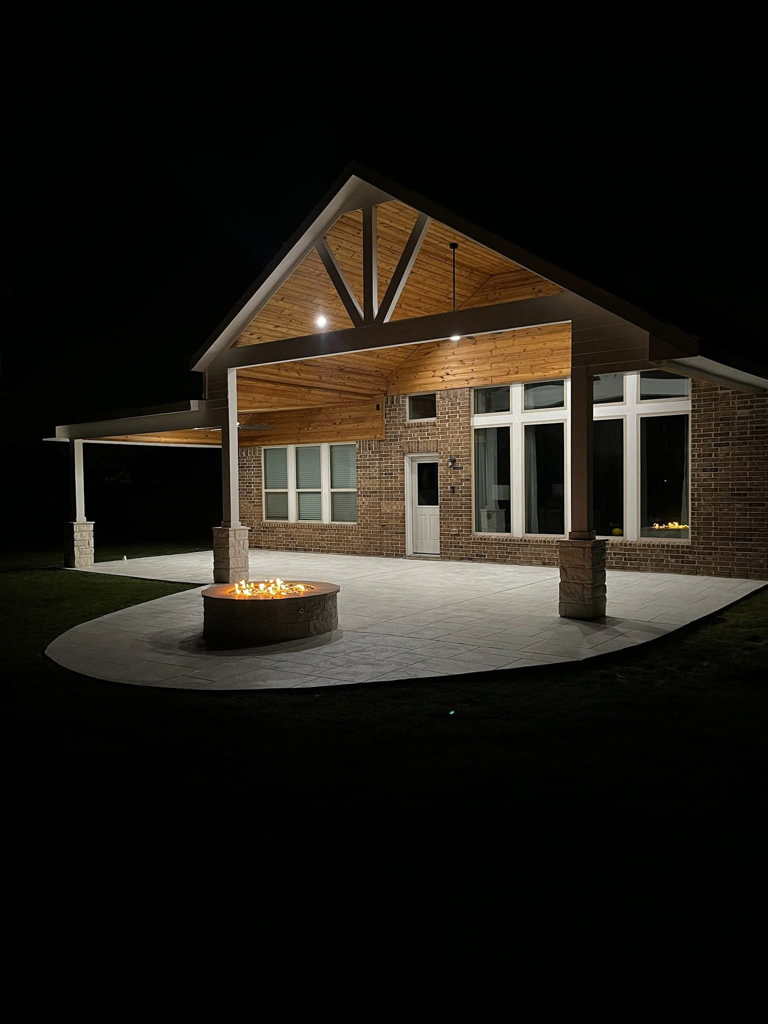 Completed covered patio with a vaulted wood ceiling, stone columns, and built-in fire pit at night